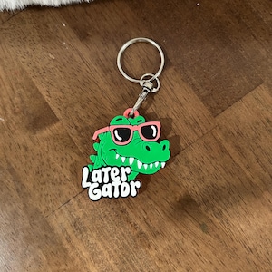 May include: A green alligator keychain featuring a cartoon character wearing pink sunglasses. The alligator has a smiling expression with visible white teeth. Below the alligator, the text "Later Gator" is written in white letters with a black outline.