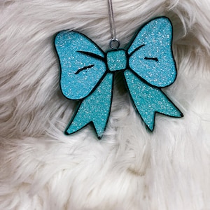 May include: A turquoise blue glitter bow ornament with a silver string hanging from it. The ornament is on a white fluffy surface.