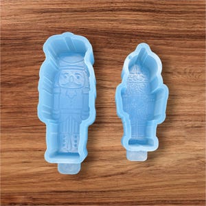May include: Two light blue silicone molds in the shape of nutcrackers. The molds are detailed with the nutcracker's uniform and facial features. The molds are on a wooden surface.