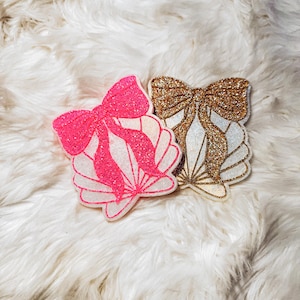 May include: Two seashell-shaped ornaments with glitter bows. One is hot pink, the other is gold. Both have white and gold accents. The ornaments are on a fluffy, white surface.