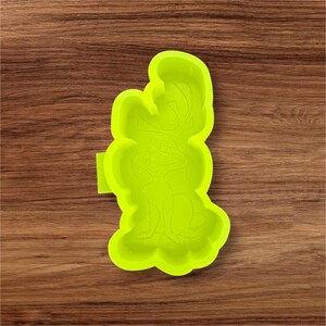 May include: A bright green silicone mold in the shape of a cartoon character. The mold features a detailed outline of the character's face, body, and ears. The mold is on a wooden surface.