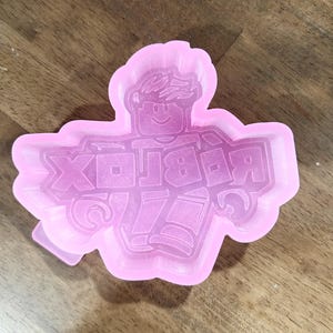 May include: A pink silicone mold featuring the Roblox logo and a cartoon character. The mold is in the shape of the Roblox logo with a smiling character in the center. The mold is designed for crafting or baking.