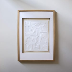 Puede incluir: Una obra de arte abstracta enmarcada. La obra presenta un diseño en relieve blanco texturizado de formas y líneas geométricas. Está enmarcada en un paspartú blanco y un marco de madera natural, creando una estética limpia y moderna. La obra está montada en una pared blanca.