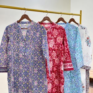 Può includere: Quattro tuniche a maniche lunghe sono appese a un'asta dorata. Le tuniche sono in vari colori e fantasie, tra cui viola con un disegno geometrico, rosso con stampe floreali, blu con stampe floreali e bianco con accenti floreali. Le tuniche hanno un davanti abbottonato.