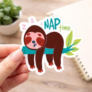 Może przedstawiać: Naklejka przedstawiająca śpiącego leniwca z kreskówki na gałęzi. Leniwiec jest brązowy z różową twarzą i czerwonymi stopami. Tekst "NAP time" jest w kolorze morskim i zielonym, z "Z" nad głową leniwca.