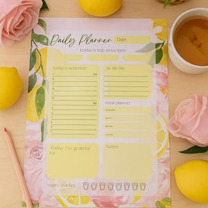 Könnte beinhalten: Ein täglicher Planer mit floralem Zitronen- und Rosendesign. Er enthält Abschnitte für die Planung, To-Do-Listen, Essensplanung und Dankbarkeit. Ein Bleistift, Zitronen und eine Tasse Tee sind ebenfalls vorhanden.