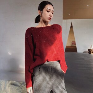 A woman wearing a red sweater with embellishments stands in a room with a neutral color palette.