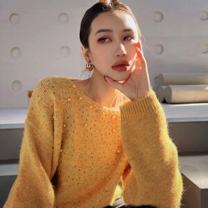 A young woman wearing a yellow sweater with sparkly details, posing with her hand on her face.