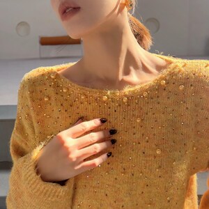 A close-up of a person wearing a yellow sweater with gold embellishments, holding their hand up to their chest.