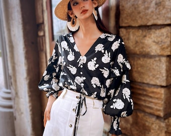 May include: A black blouse with white floral print, featuring a V-neck and long, gathered sleeves. The blouse is paired with a white skirt. The outfit is accessorized with a straw hat and earrings. The overall style is feminine and summery.