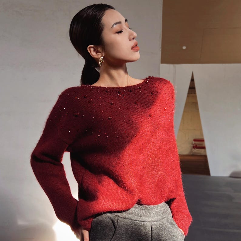 A woman wearing a red sweater with embellishments stands in a room with a neutral background.