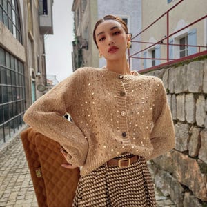 Puede incluir: Un cárdigan beige con lentejuelas y cuello redondo. El suéter abotonado tiene mangas largas. El atuendo incluye un cinturón marrón y una falda estampada marrón y blanca. Se ve un bolso acolchado marrón.