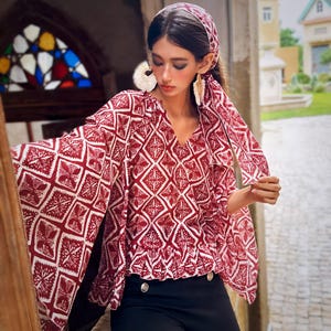 May include: A burgundy and white patterned blouse with a V-neck and flared sleeves. The blouse is paired with black pants and a matching headscarf. The outfit is accessorized with large earrings.