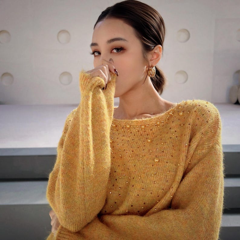 A woman wearing a yellow sweater with sparkly details, posing with her hand on her face.