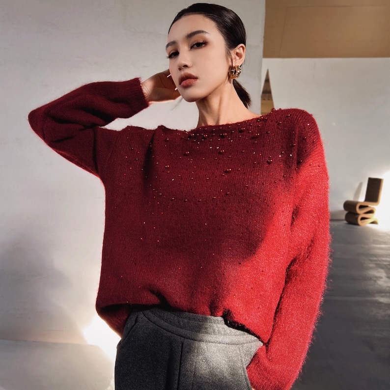A woman wearing a red sweater with sparkly embellishments poses in a room with a wooden chair visible in the background.
