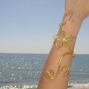 May include: Gold-toned arm jewelry featuring a floral bracelet with a pearl center and two swirling arm cuffs. The jewelry is displayed against a backdrop of a beach and ocean under a clear blue sky.