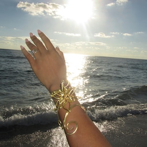 May include: A gold-colored arm cuff with a floral design and swirling accents. The cuff is worn on an arm with a ring on a finger. The background shows a beach with waves and the sun shining brightly.