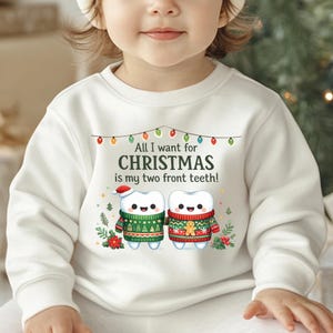 May include: White sweatshirt with the text "All I want for Christmas is my two front teeth!" and a graphic of two cartoon teeth wearing Christmas sweaters. One tooth has a Santa hat. The design includes Christmas lights and festive greenery.