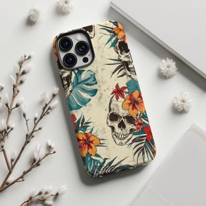 以下が含まれることがあります： スカルと花の模様の携帯電話ケース。ケースはクリーム色の背景に、スカル、熱帯の花、葉のイラストがオレンジ、赤、ティール色で描かれています。ケースのカメラの周りには黒い縁があります。