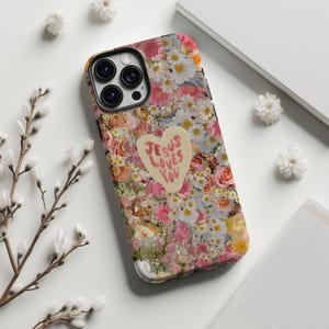 May include: A floral phone case featuring a heart-shaped design with the text "JESUS LOVES YOU." The case is covered in a mix of pink, white, and yellow flowers, including daisies and roses. The phone case has a black camera lens.