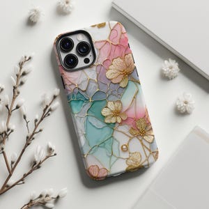 Può includere: Custodia per smartphone con design floreale e dettagli dorati. La custodia ha una base bianca con motivi rosa, turchese e grigi ad acquerello. Fiori color oro e un contorno dorato completano il design.