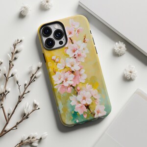 以下が含まれることがあります： ピンクと白の花柄が特徴の黄色の携帯電話ケース。ケースには黒いカメラモジュールと黒いサイドボタンが付いています。背景は、黄色、緑、青の色合いの水彩効果があります。