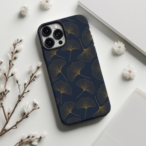 May include: A navy blue phone case featuring a repeating pattern of gold ginkgo leaf outlines. The case has a black border around the camera and a glossy finish. The phone case is on a white surface with floral accents.