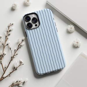 May include: A light blue phone case with vertical navy blue stripes. The case has a black camera lens protector and is surrounded by white flowers and a white background.