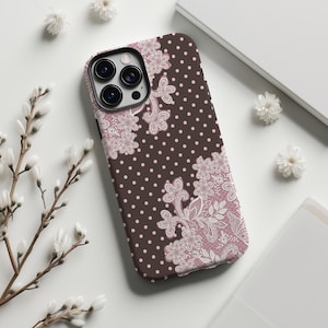 以下が含まれることがあります： 茶色の水玉模様と白とピンクの花柄レースのデザインが施された携帯電話ケース。ケースにはカメラ用の切り抜きがあり、白い花と白い背景に囲まれています。