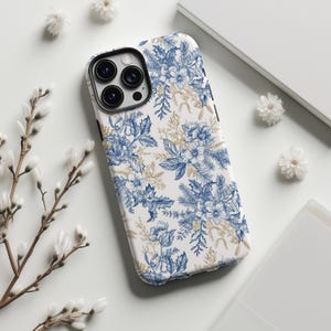 以下が含まれることがあります： 白地に青い花柄とゴールドのアクセントが施された携帯電話ケース。ケースは、さまざまな花や葉を使った詳細な植物のデザインが特徴です。ケースはスマートフォンに合うように設計されています。