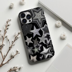 以下が含まれることがあります： 黒い携帯電話ケース、星柄。ケースは、シルバー、グリッター、メタリック仕上げのさまざまな星のデザインが特徴です。星は黒い背景に配置されています。携帯電話ケースには、カメラの周りに黒い縁があります。