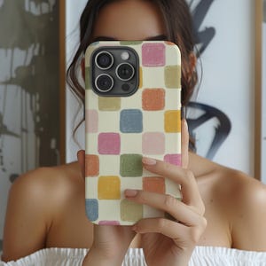 May include: A phone case with a colorful, checkered pattern. The case features a cream-colored background with squares in shades of pink, blue, green, orange, and yellow. The phone case is held up in front of a person's face.