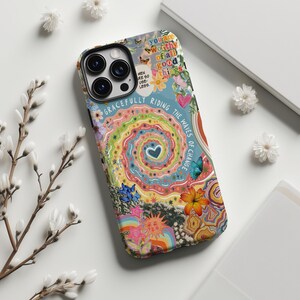 以下が含まれることがあります： 花と蝶のデザインが施されたカラフルな携帯電話ケース。ケースの中央にはハートの螺旋模様があります。「You are worthy of all good things」と「Gracefully riding the waves of change」というテキストが含まれています。