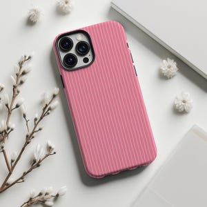 May include: A pink phone case with a vertical white pinstripe pattern. The case has a black camera lens surround and black side buttons. The phone case is surrounded by white flowers and a white book.