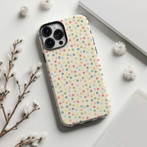 May include: A phone case with a cream-colored background and a pattern of colorful polka dots. The case has a black border and a camera cutout. The phone case is lying on a white surface with white flowers.