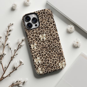 May include: A phone case with a leopard print design, featuring brown spots on a tan background. The case is adorned with illustrations of butterflies in shades of brown and white. The phone case is shown on a white surface.