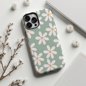 May include: A phone case featuring a daisy pattern. The case has a pale green background with white daisies and yellow centres. The phone case is on a white surface with white flowers.