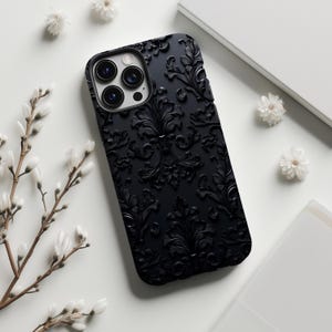 May include: Black phone case with a raised floral pattern. The case has a matte finish and features a camera cutout. White flowers and a white book are in the background.