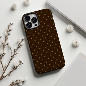 May include: A brown phone case with a repeating gold fleur-de-lis pattern. The case has a camera cutout and is surrounded by white flowers and a white background.