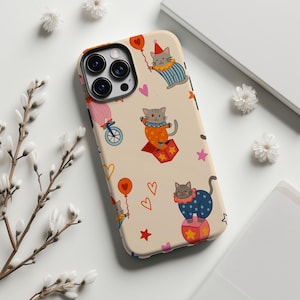 以下が含まれることがあります： ベージュ色の携帯電話ケース。サーカスの猫をモチーフにした、遊び心のあるデザインです。ピエロの衣装を着た猫、風船、ハート、星が描かれています。ケースにはカメラ用の切り抜きと保護バンパーが付いています。