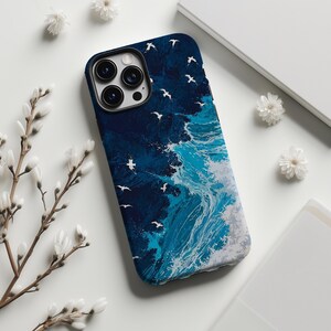 Può includere: Custodia per telefono con un design a tema marino, raffigurante gabbiani bianchi che volano sopra le onde che si infrangono. La custodia ha un bordo blu scuro e una vivace scena oceanica blu, bianca e grigia. Presentata su una superficie bianca con fiori decorativi.