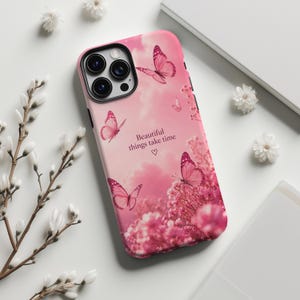 以下が含まれることがあります： 曇り空を背景に、蝶とピンクの花がプリントされたピンクのスマートフォンケース。中央には「Beautiful things take time」という文字がプリントされており、その下に小さなハートのアイコンが配置されています。