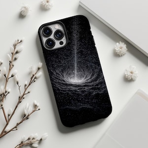 May include: Black phone case featuring a white and gray abstract design resembling a cosmic event. The case has a glossy finish and a camera cutout. White flowers and a white notebook are in the background.