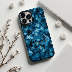 以下が含まれることがあります： 小さな青い花と黄色の中心が暗い背景に描かれた、花柄のスマートフォンケース。ケースには黒い縁とカメラの切り抜きがあります。白い花と白い表面がケースを囲んでいます。