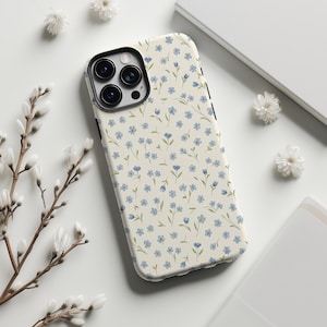 Puede incluir: Funda de teléfono con fondo crema y un estampado de pequeñas flores azules y tallos verdes. La funda tiene un borde negro alrededor de las lentes de la cámara y un borde lateral negro. Flores y ramas blancas rodean la funda.