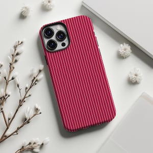 May include: A red phone case with a vertical white pinstripe pattern. The case has a black border and a camera cutout. The phone case is lying on a white surface with white flowers and a book.