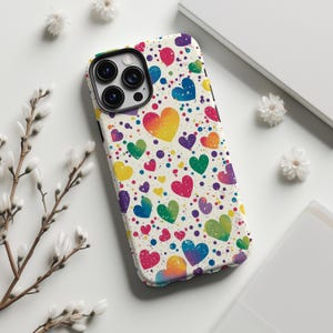 以下が含まれることがあります： カラフルなハート模様の携帯電話ケース。赤、黄、緑、青など、さまざまなサイズと色のハートが、カラフルな水玉模様の白い背景に描かれています。ケースには、カメラレンズの周りに黒い縁取りがあります。