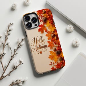 Può includere: Una custodia per smartphone con sfondo color crema che riporta la scritta Hello Autumn in corsivo marrone. Il lato destro è decorato con un motivo vivace di foglie d'acero arancioni e rosse, ghiande marroni, pigne e piccole bacche rosse.