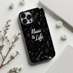 以下が含まれることがあります： 黒い携帯電話ケース。白い音符と楽器のパターン。白い文字で「Music is Life」と書かれています。白い背景に白い花が添えられています。