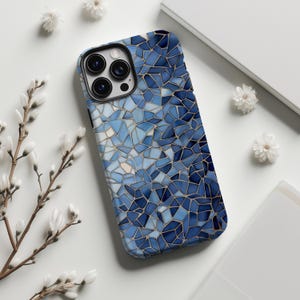 Pode incluir: Uma capa de smartphone com um design de mosaico em tons de azul. A capa apresenta um padrão de formas irregulares, criando uma aparência texturizada. A capa tem um módulo de câmera preto. Flores brancas e um fundo branco circundam a capa.
